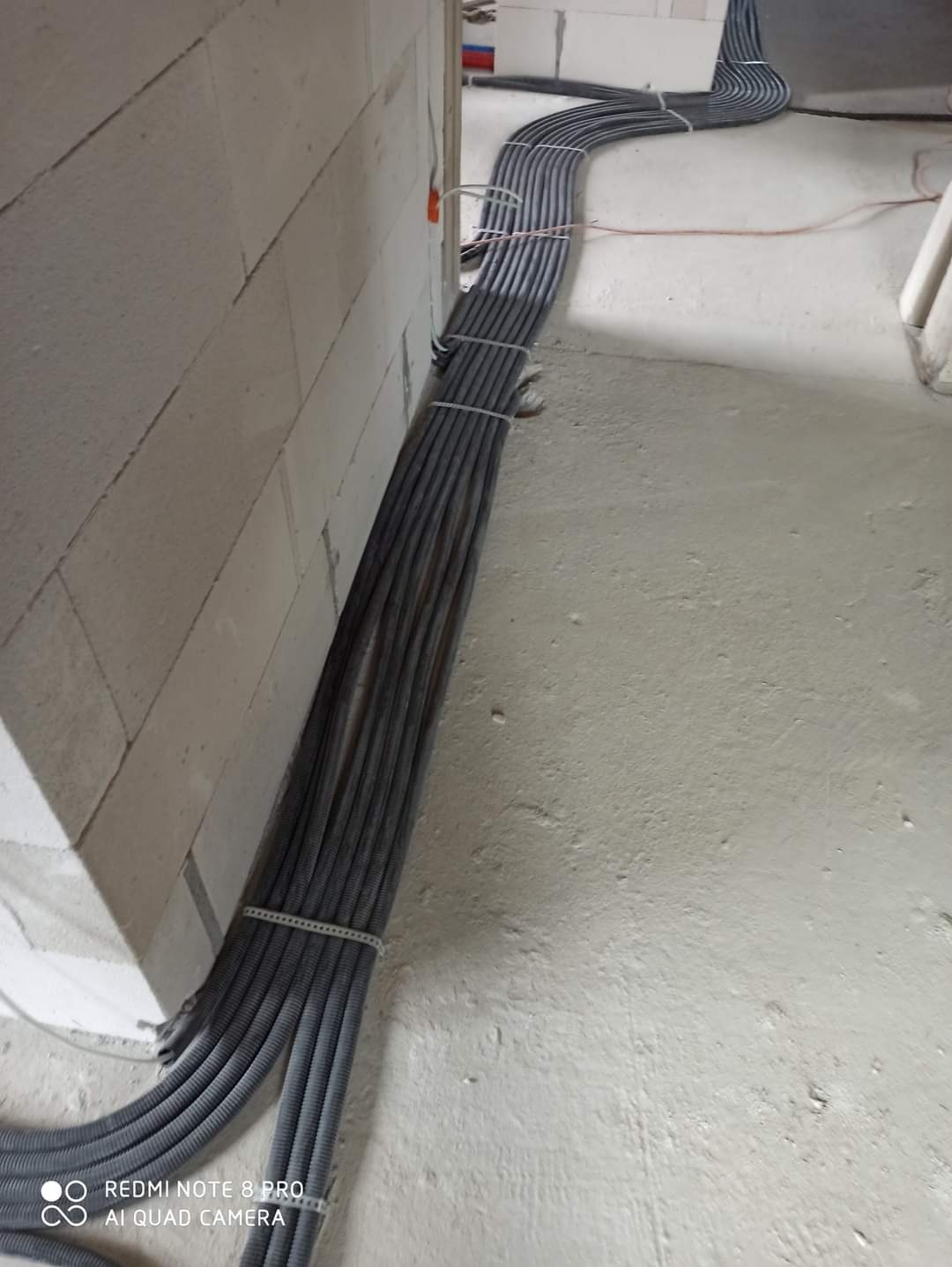 Szereg szarych rur karbowanych, przymocowanych do ściany z białych bloczków, układających się wzdłuż podłogi w surowym stanie. Widoczne mocowania i fragment instalacji.