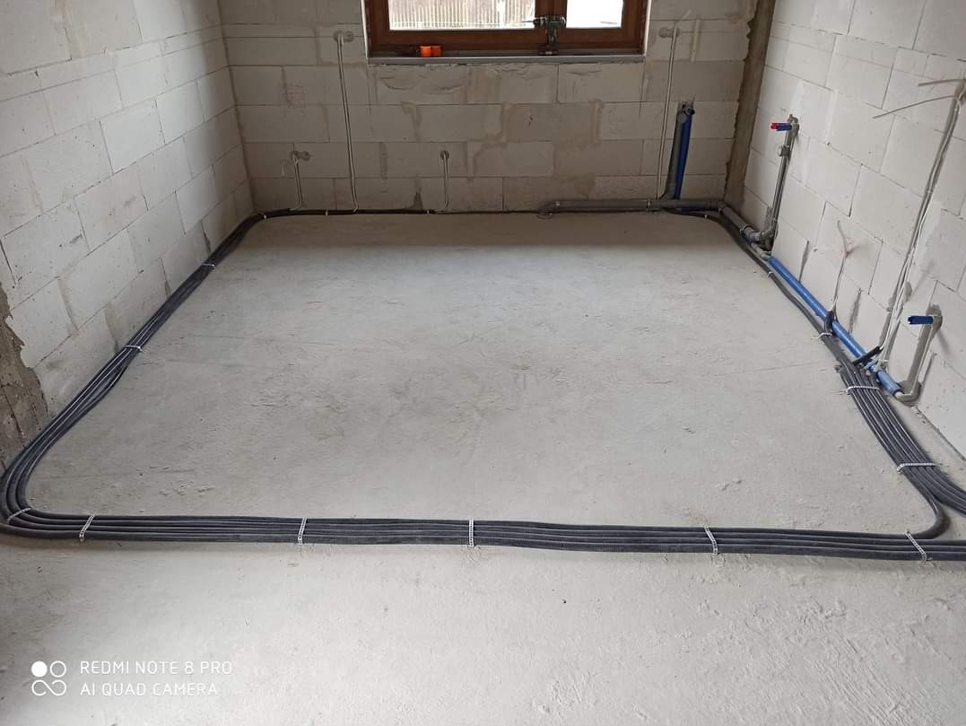 Szara podłoga w surowym wnętrzu z instalacją elektryczną w listwach przypodłogowych i rurami wodnymi przy ścianach z betonu komórkowego.