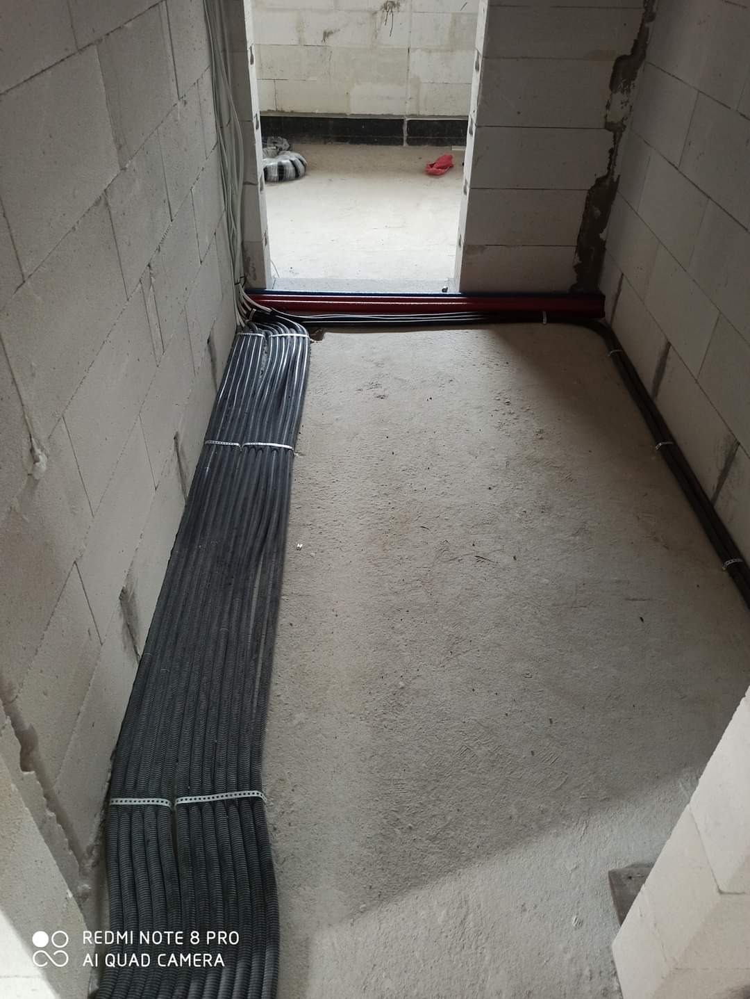 Wiązka kabli w peszlu, ułożona wzdłuż ściany z betonu komórkowego, z widocznymi mocowaniami. Czerwone rury instalacyjne w tle, surowy stan wykończenia.