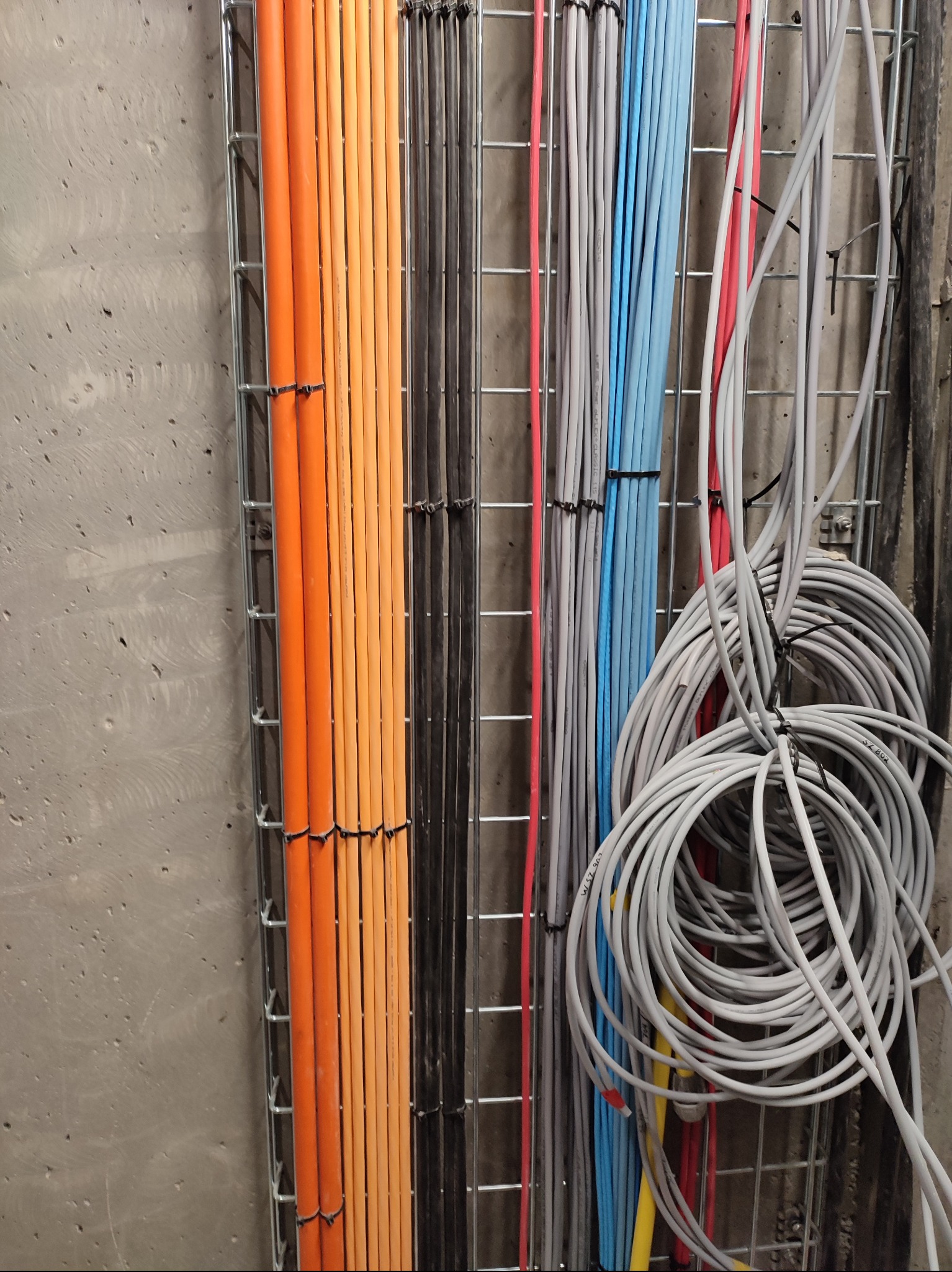 Różnokolorowe kable elektryczne ułożone w metalowej siatce na szarej ścianie. Widoczne pomarańczowe, czarne, szare i niebieskie przewody. Część kabli zwinięta w pętle.
