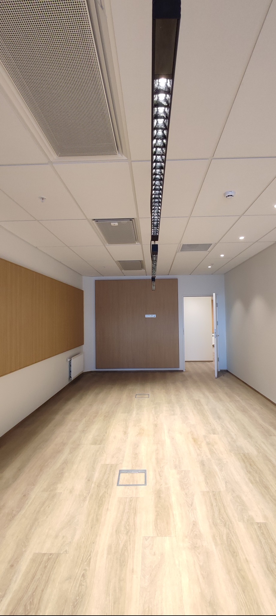 Nowoczesne oświetlenie biurowe w Ełku: długa lampa sufitowa LED w jasnym pomieszczeniu z drewnianymi panelami i podłogą. Widoczne kratki wentylacyjne.