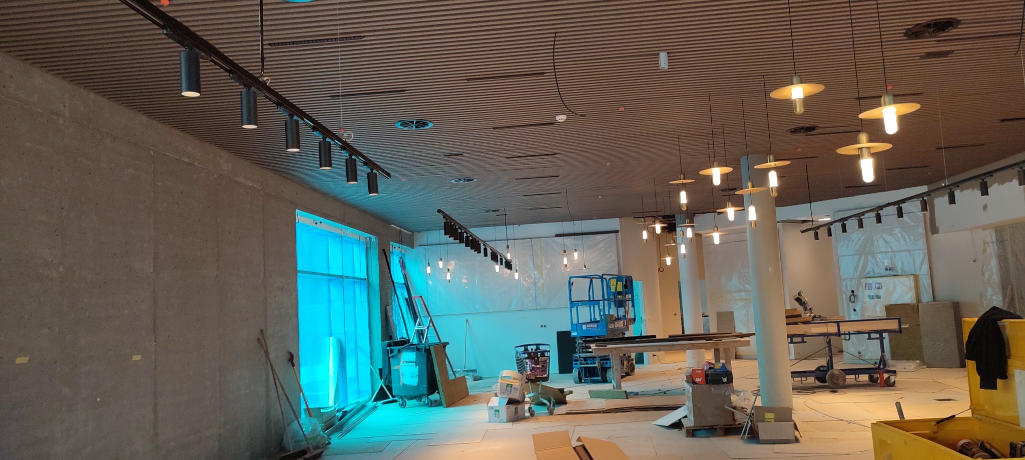 Montaż oświetlenia w nowym lokalu: czarne reflektory szynowe i wiszące lampy z metalowymi kloszami. Widoczny podnośnik i materiały budowlane.