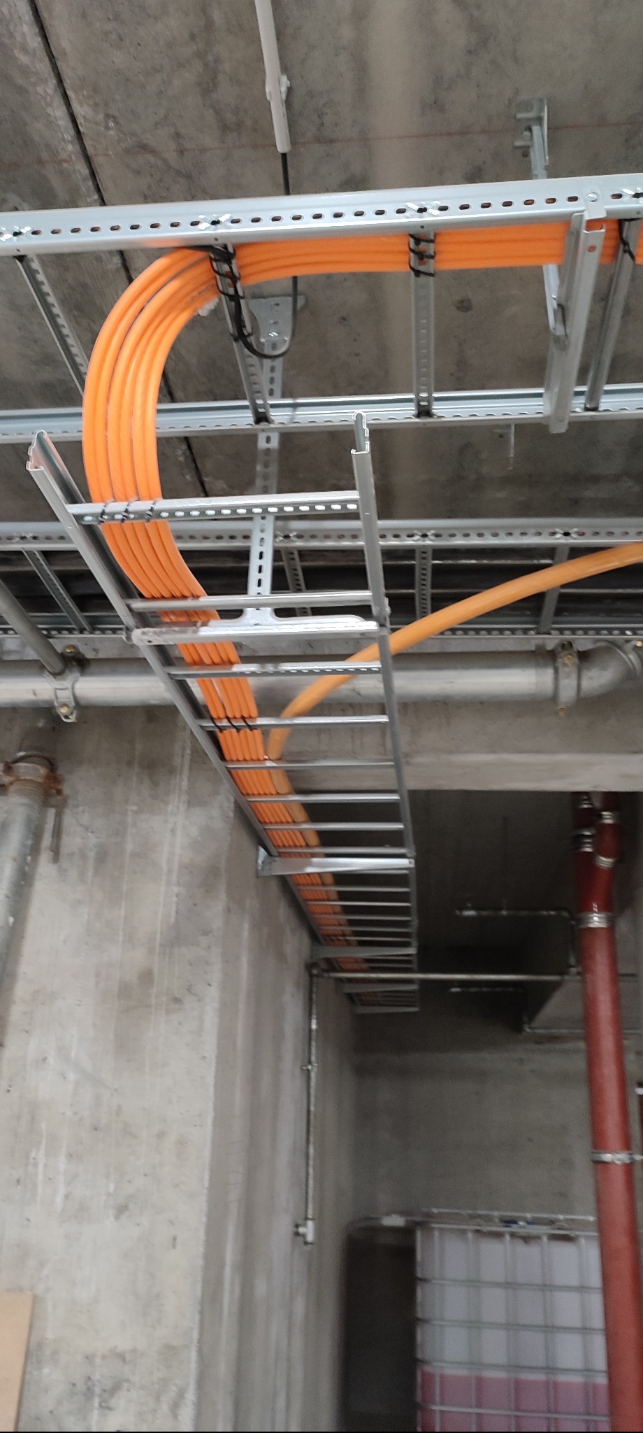 Pomarańczowe kable ułożone w metalowej korycie pod sufitem, zabezpieczone czarnymi opaskami. Widoczne rury i betonowe ściany w tle. Instalacja elektryczna w surowym stanie.