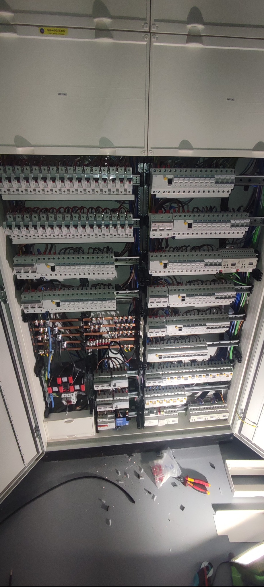 Otwarta szafa elektryczna z licznymi bezpiecznikami, przewodami i elementami sterującymi. Widoczne narzędzia i pozostałości po montażu na podłodze.