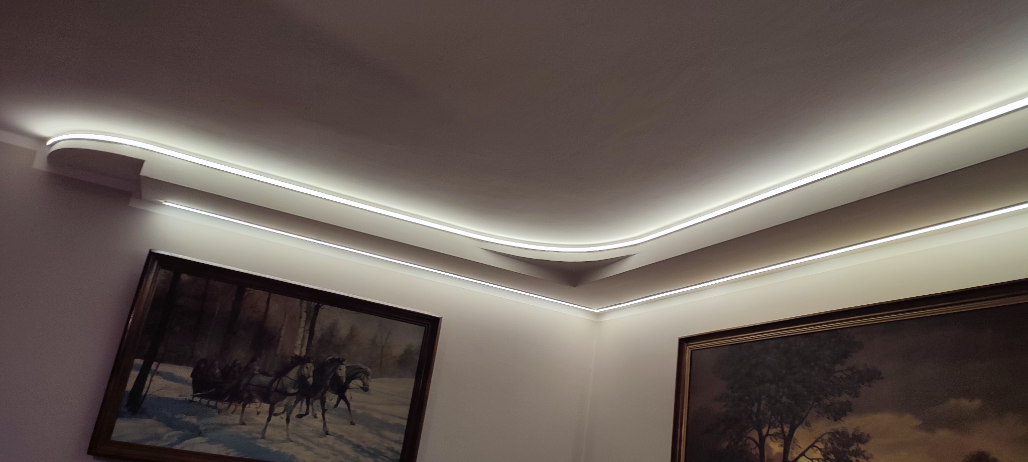 Nowoczesne oświetlenie LED wzdłuż sufitu, podkreślające dekoracyjne listwy i obrazy na ścianach. Subtelne, białe światło tworzy elegancki nastrój we wnętrzu.