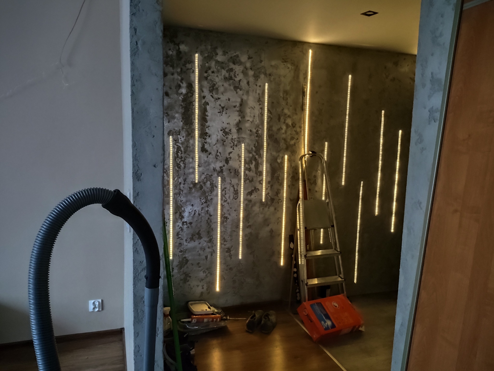 Ściana z dekoracyjnym oświetleniem LED w postaci pionowych linii. Widoczna drabina, narzędzia i wąż od odkurzacza. Surowy beton na ścianie.