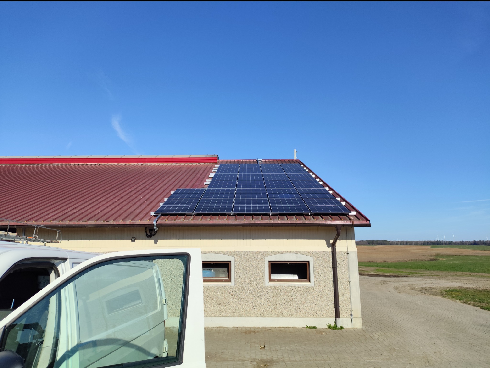 Panele słoneczne na dachu budynku z czerwoną dachówką, widok z boku z częściowo widocznym białym samochodem dostawczym. Błękitne niebo w tle.