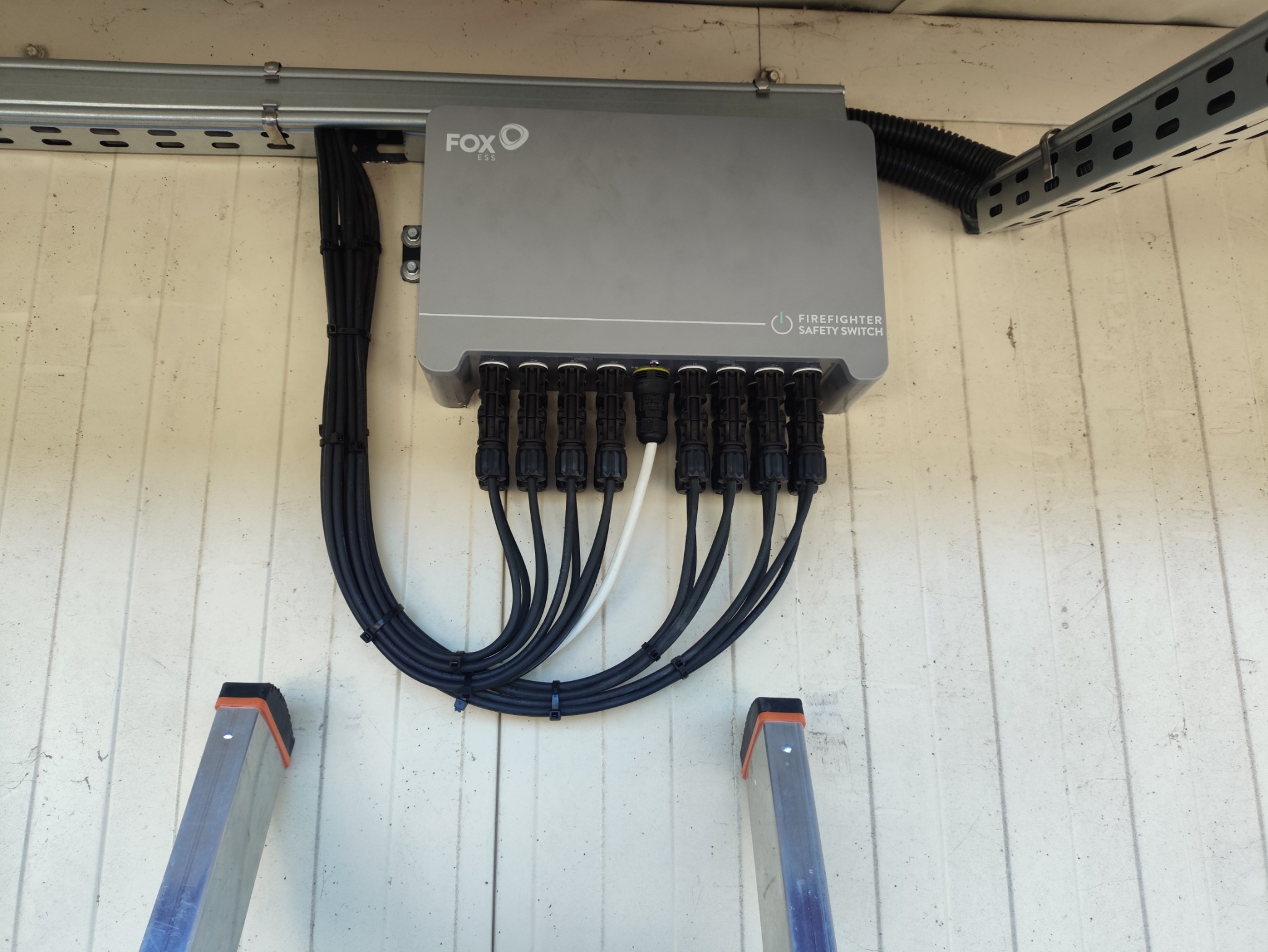 Szary moduł FOX ESS z podłączonymi czarnymi kablami, zamontowany na ścianie. Widoczne fragmenty drabiny. Urządzenie z napisem 'FIREFIGHTER SAFETY SWITCH'.