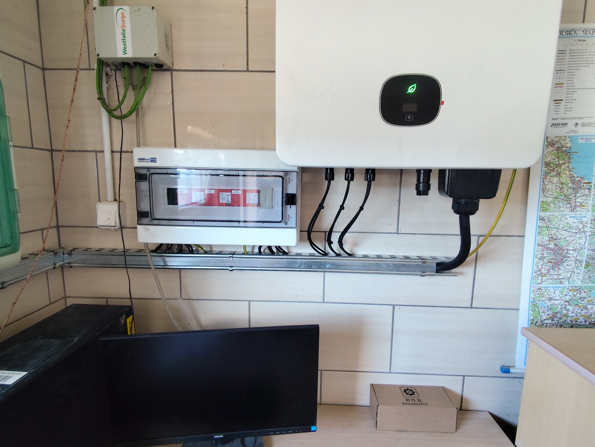 Biała skrzynka z zielonym symbolem liścia, pod nią rozdzielnia elektryczna z przewodami. Na ścianie mapa. Elementy instalacji elektrycznej w pomieszczeniu.