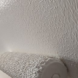 ORDEX SPÓŁKA Z OGRANICZONĄ ODPOWIEDZIALNOŚCIĄ - Malowanie ściany wałkiem z wyraźną fakturą, świeżo nałożona biała farba, zbliżenie na strukturę. Widoczny fragment podłogi zabezpieczony folią malarską.