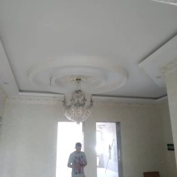 ORDEX SPÓŁKA Z OGRANICZONĄ ODPOWIEDZIALNOŚCIĄ - Wykończony sufit z dekoracyjnym gipsem i żyrandolem w folii ochronnej. Ściany pomalowane na jasny kolor. Widoczny fragment pomieszczenia w trakcie prac wykończeniowych.