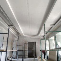 ORDEX SPÓŁKA Z OGRANICZONĄ ODPOWIEDZIALNOŚCIĄ - Wykończony sufit z płyt karton-gips z dekoracyjnymi elementami, widok z dołu, rusztowania w pomieszczeniu, osoba w tle. Detal wykończenia wnętrza.