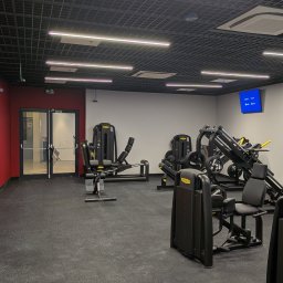 ORDEX SPÓŁKA Z OGRANICZONĄ ODPOWIEDZIALNOŚCIĄ - Nowoczesna siłownia z czarną podłogą i sufitem rastrowym, oświetlona ledami. Widoczne profesjonalne maszyny do ćwiczeń Technogym i czerwona ściana.