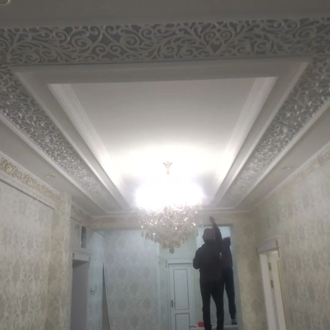 Wykończony sufit z dekoracyjnymi elementami sztukaterii i oświetleniem. Dwóch pracowników na drabinie podczas prac wykończeniowych w pomieszczeniu z tapetami.