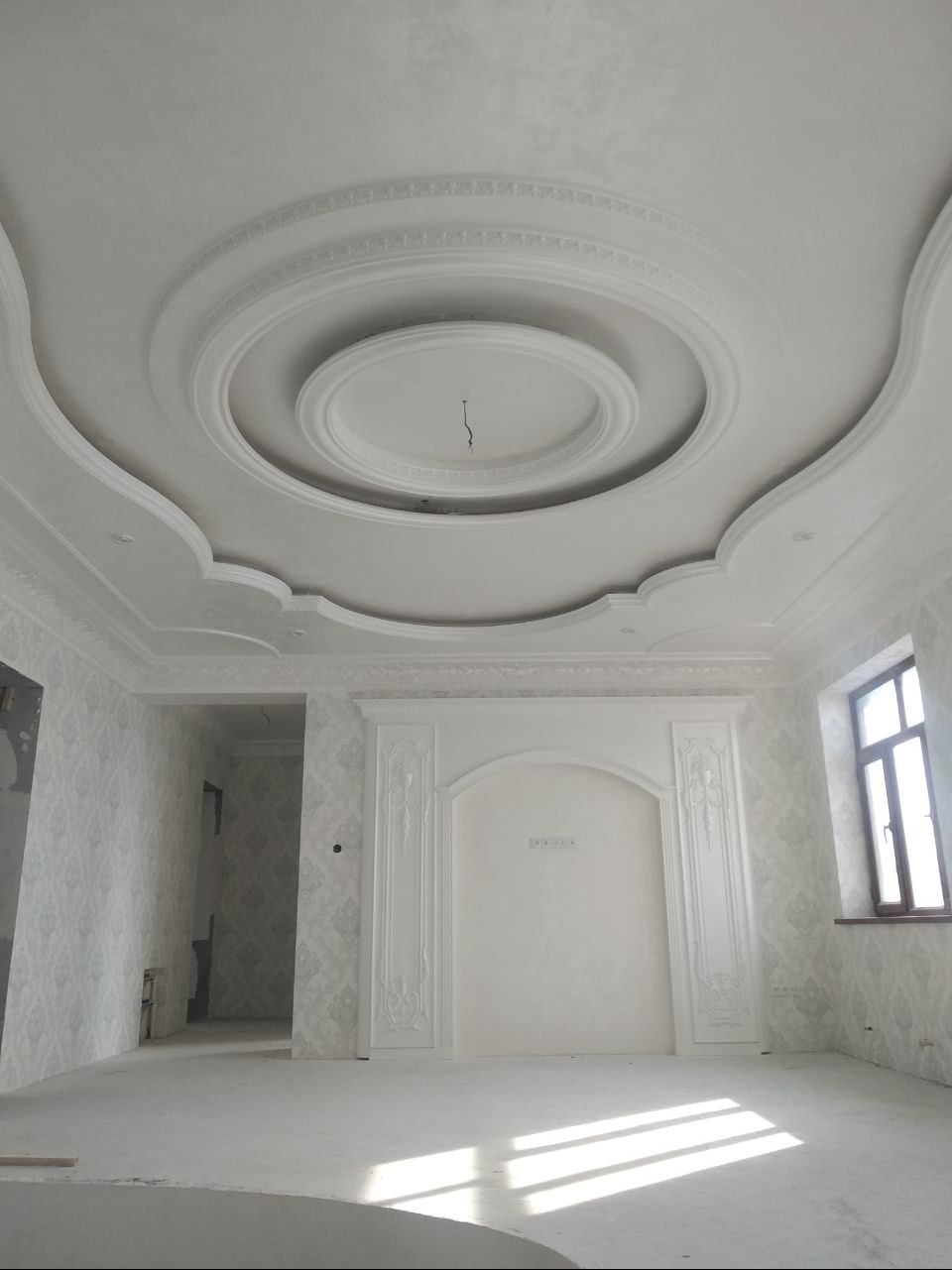 Eleganckie wykończenie wnętrza z dekoracyjnym sufitem i sztukaterią ścienną w jasnej tonacji. Widoczne detale wykończeniowe i okno z drewnianą ramą.