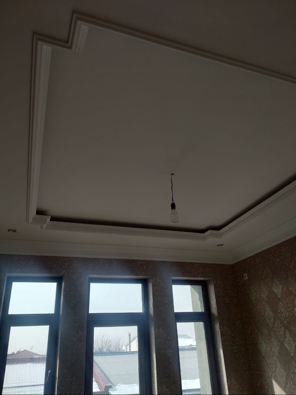 Wykończony sufit z dekoracyjnymi listwami i oświetleniem punktowym, widok z dołu na trzy okna z widokiem na zewnątrz. Tapeta w tle.