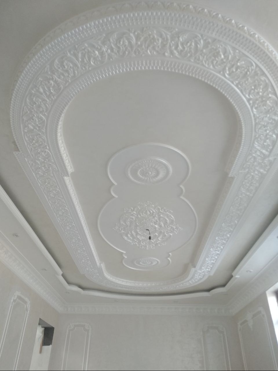 Dekoracyjny sufit z gipsowymi zdobieniami w kształcie rozet i ornamentów, wykończony w jasnej tonacji, widok z dołu, fragment ściany z listwami.