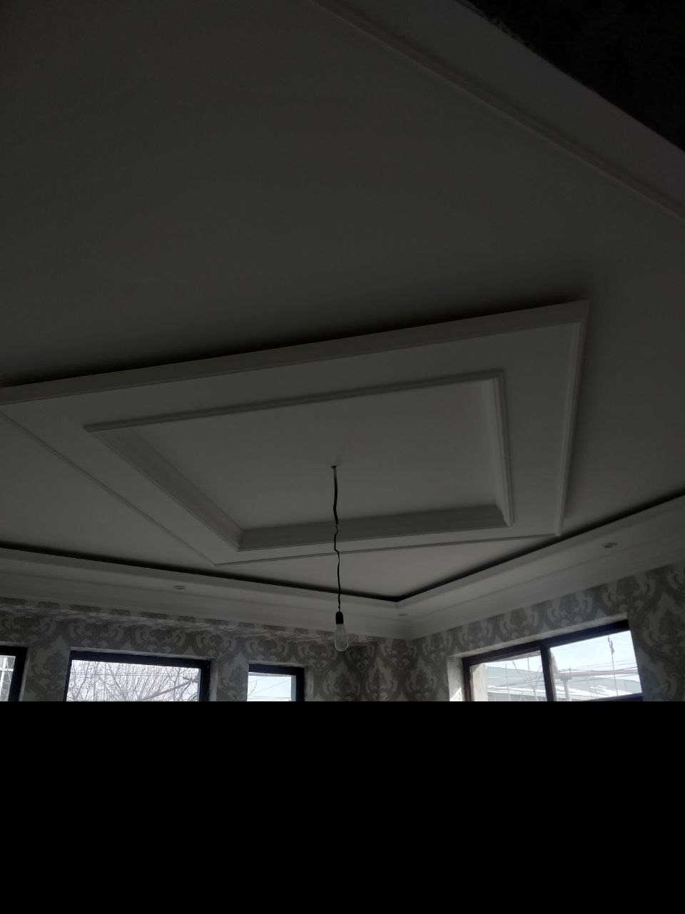 Wykończony sufit z dekoracyjnym obramowaniem i centralnym wgłębieniem, widok z dołu, tapeta w tle, surowa żarówka zwisająca z sufitu.