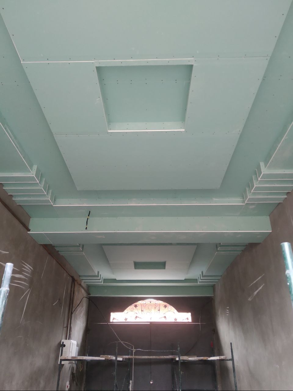 Sufit z płyt kartonowo-gipsowych z dekoracyjnymi wgłębieniami w kształcie kwadratów, widok z dołu, surowe ściany w tle, etap prac wykończeniowych.