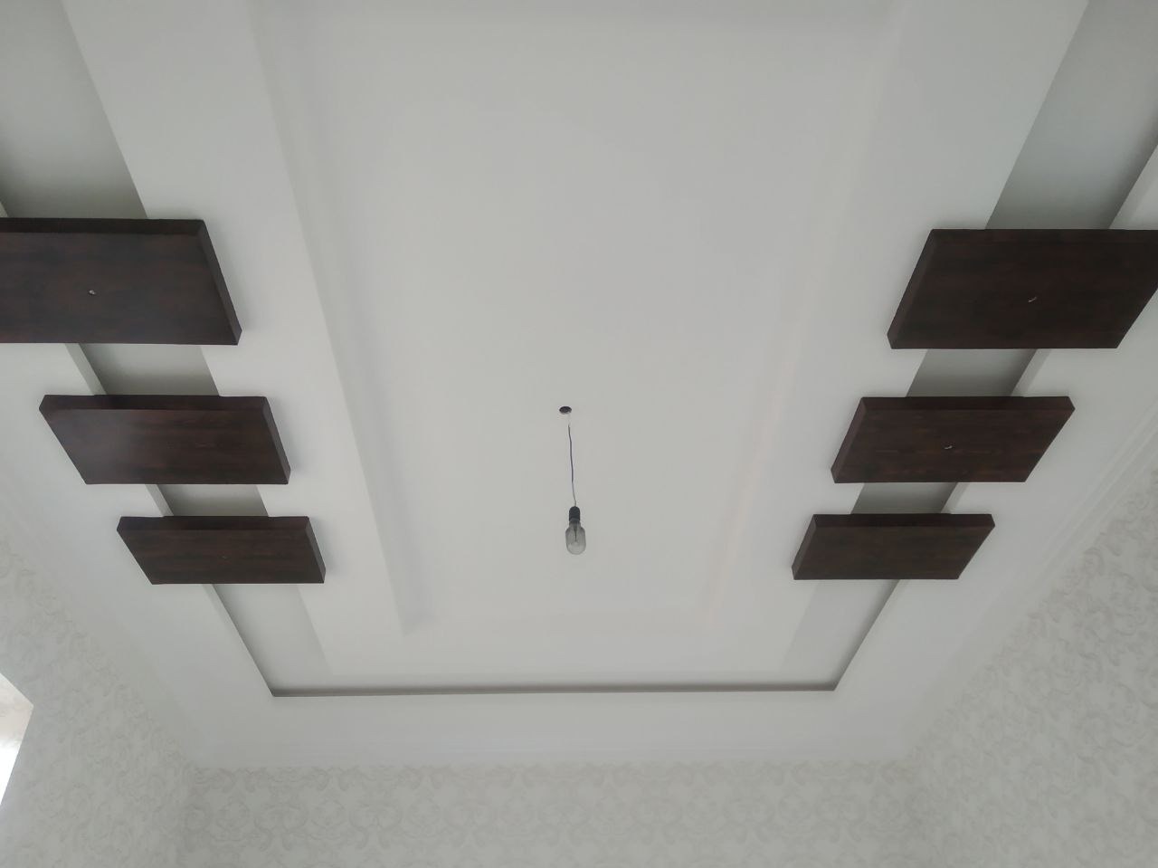 Biały sufit podwieszany z drewnianymi elementami dekoracyjnymi i wiszącą żarówką. Geometryczny wzór, symetryczny układ, delikatna tapeta w tle.