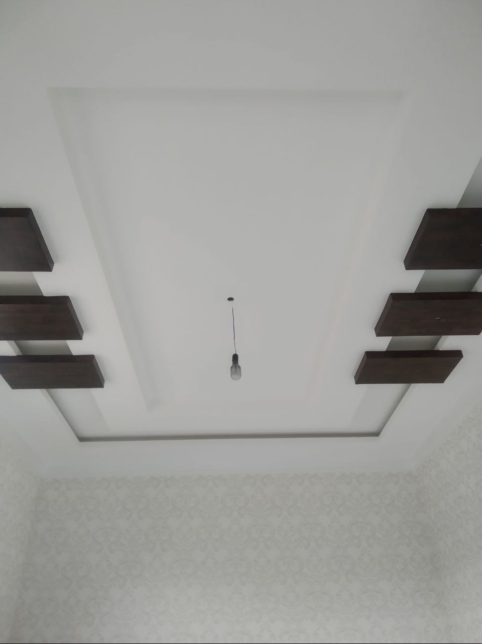 Nowoczesny sufit podwieszany z drewnianymi elementami dekoracyjnymi i minimalistyczną lampą wiszącą na tle ściany z delikatnym wzorem.