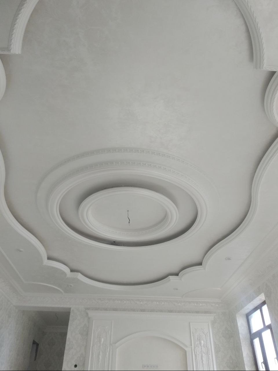Biały sufit z dekoracyjnymi sztukateriami i gładzią, oświetlony naturalnym światłem z okna. Widoczne detale wykończenia i ornamentyka.