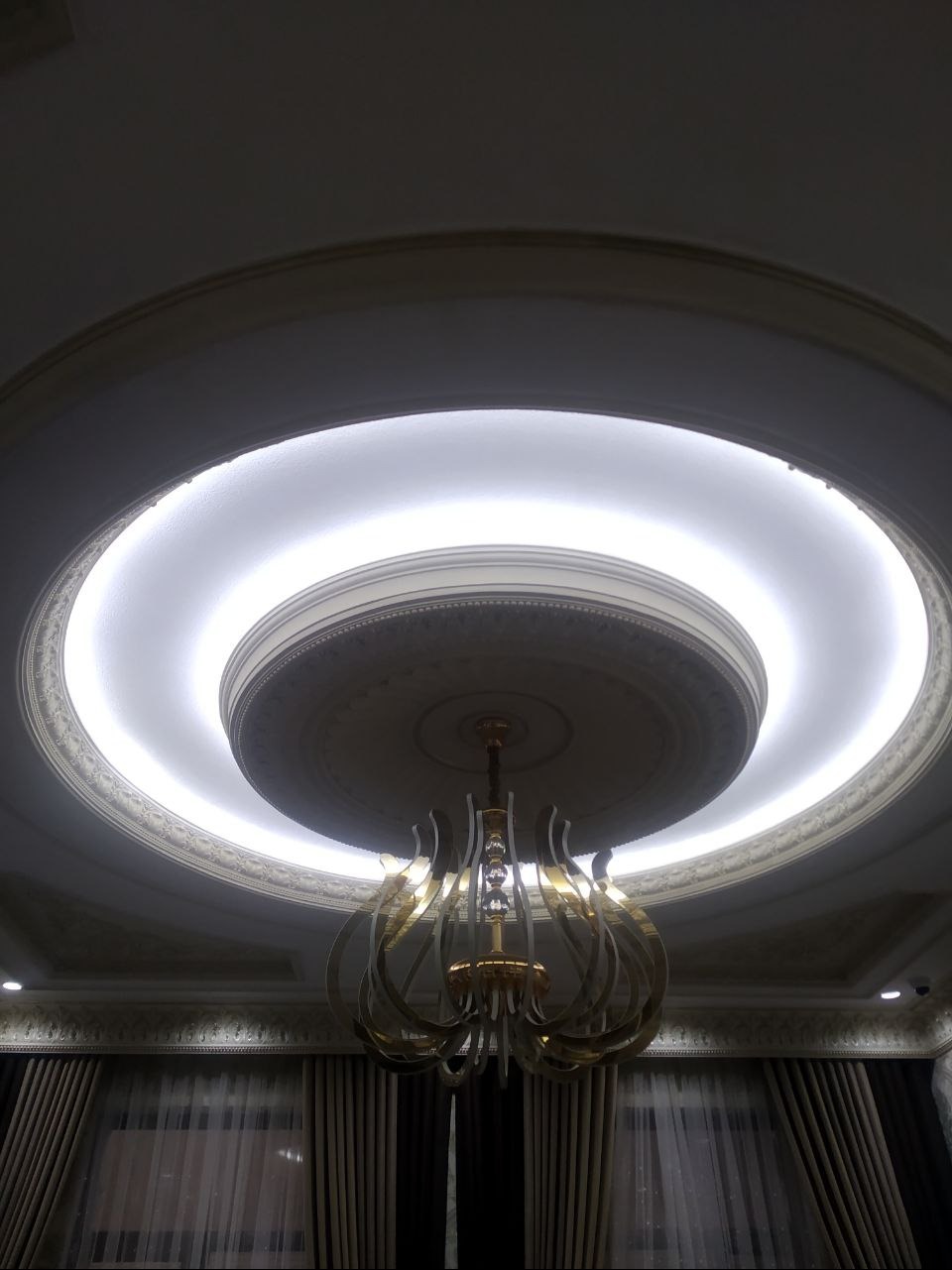 Okrągły sufit z dekoracyjnym oświetleniem LED i ozdobnym żyrandolem w stylu glamour. Widoczne detale sztukaterii i zasłony w tle.