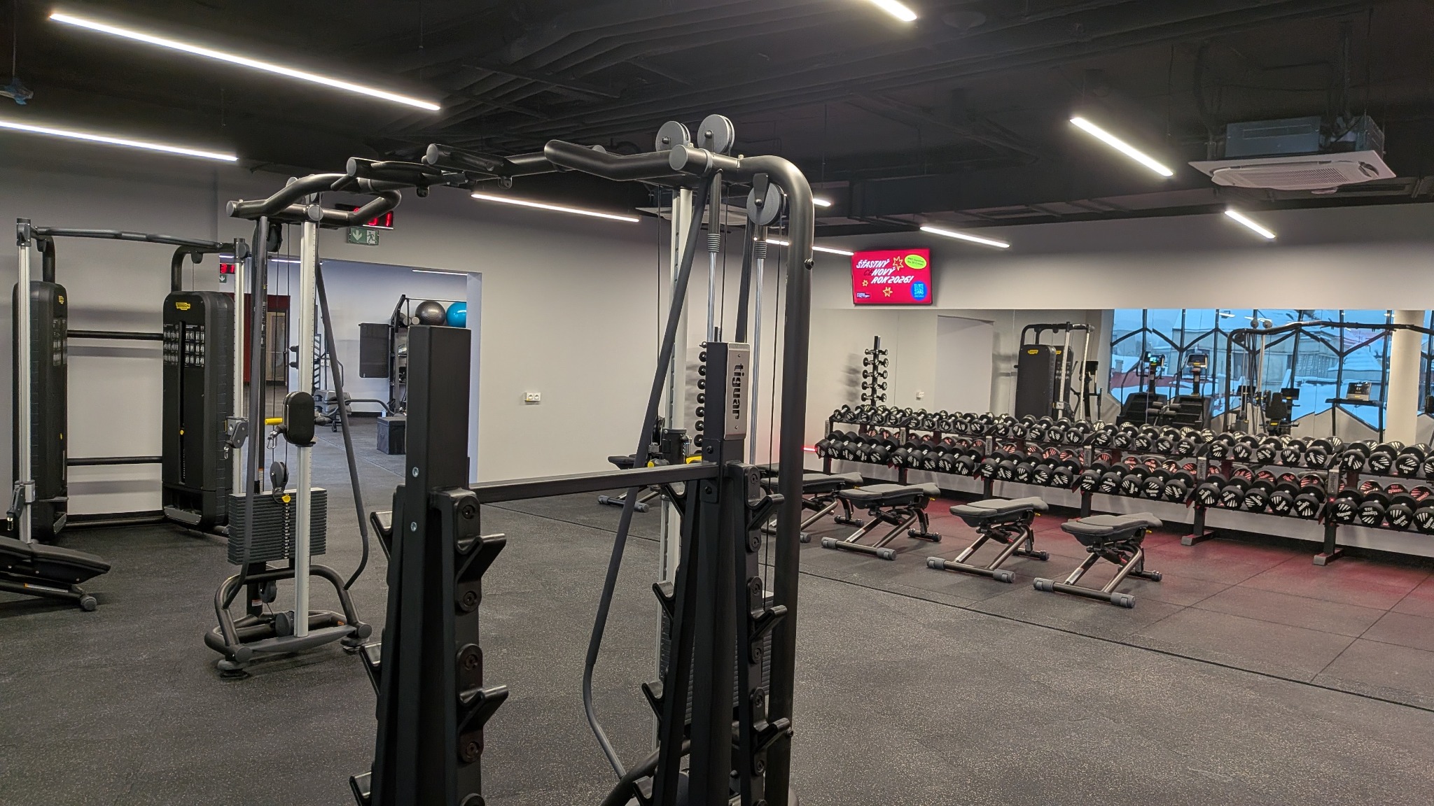 Nowoczesna siłownia z profesjonalnym sprzętem do ćwiczeń siłowych, hantlami i czarną podłogą. Widoczne oświetlenie LED i wentylacja sufitowa.