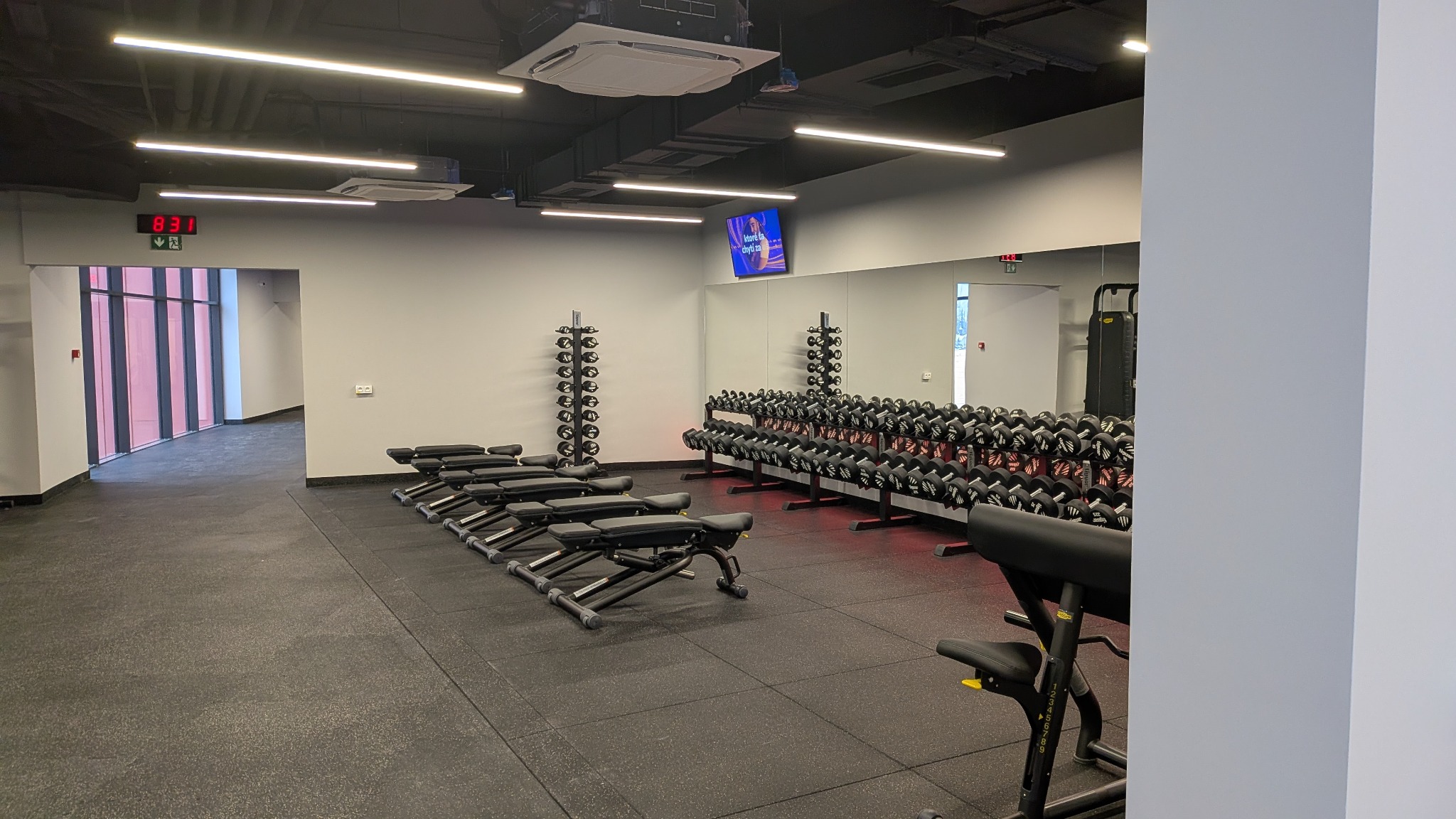 Nowoczesna siłownia z rzędami ławek do ćwiczeń i stojakami z hantlami, oświetlona ledowymi lampami sufitowymi. Widoczne lustra i telewizor na ścianie.