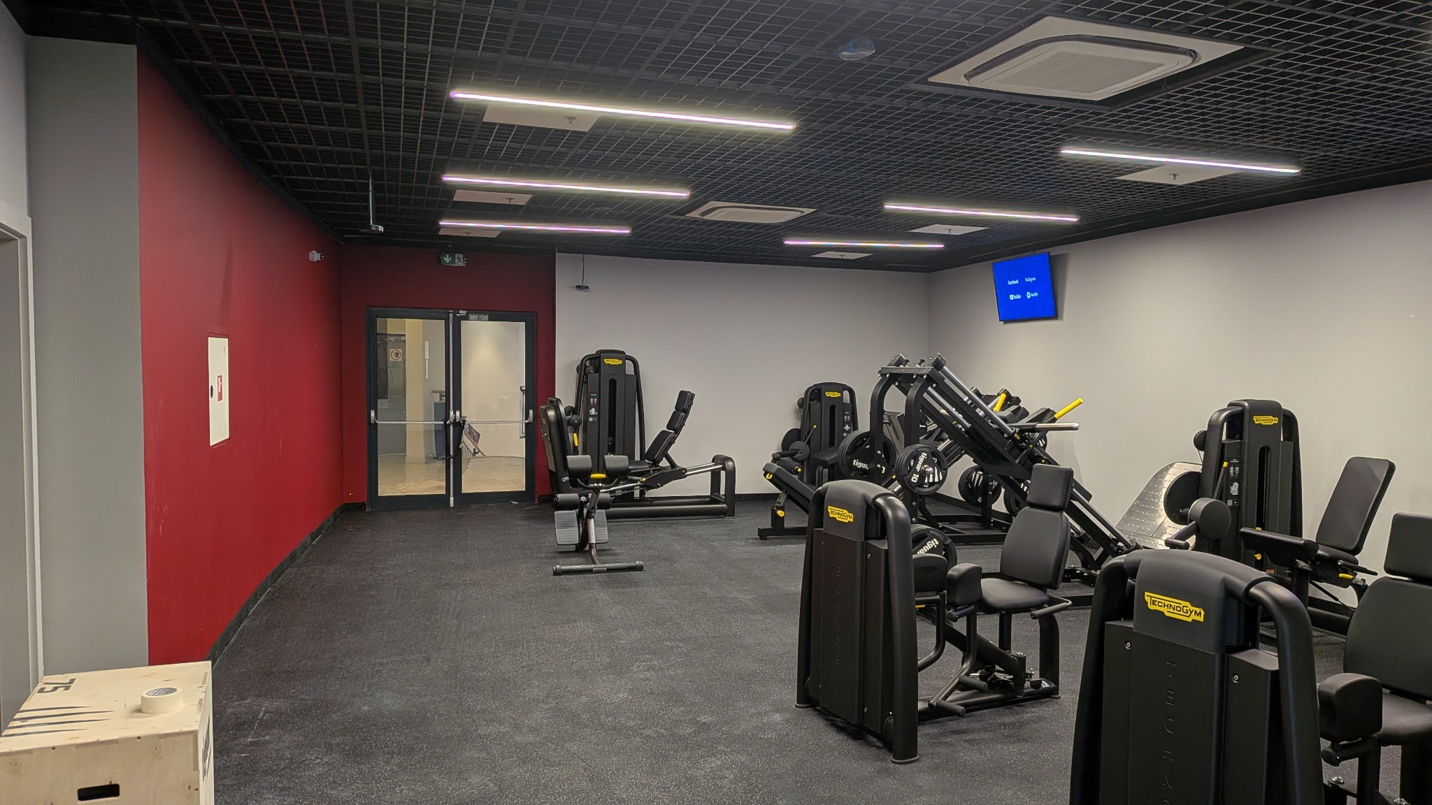 Nowoczesna siłownia z czarną podłogą i sufitem rastrowym, oświetlona ledami. Widoczne profesjonalne maszyny do ćwiczeń Technogym i czerwona ściana.