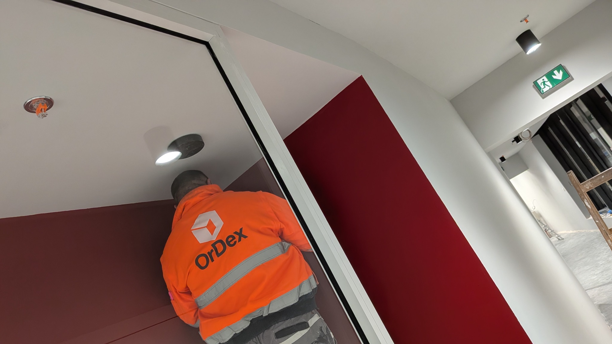 Pracownik w pomarańczowej kurtce maluje ścianę na bordowo w biurze. Widoczny sufit z oświetleniem i systemem przeciwpożarowym. Ukończony fragment ściany.