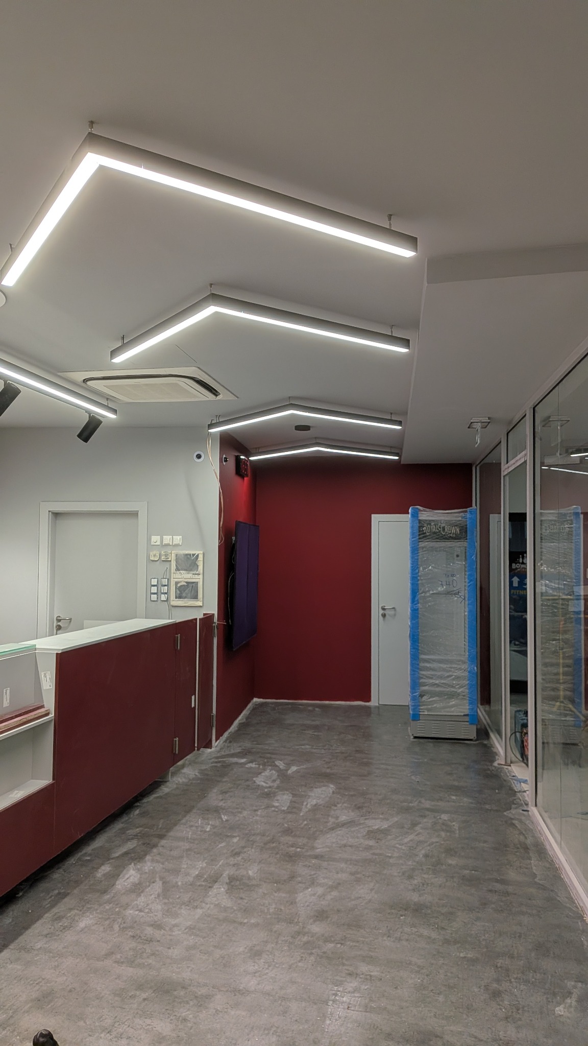 Wnętrze biura w trakcie prac wykończeniowych: surowa podłoga, ściany w kolorze bordo, nowoczesne oświetlenie LED i przeszklone ściany działowe. Widoczny fragment lady recepcyjnej.