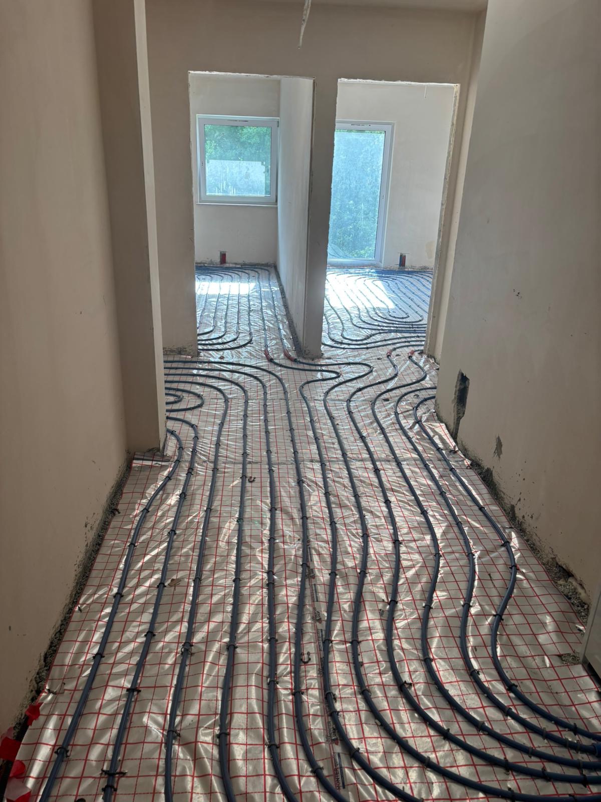 Ułożone rury ogrzewania podłogowego na folii z siatką w surowym wnętrzu. Widok z korytarza na dwa pomieszczenia z oknami. Ściany przygotowane do dalszych prac.