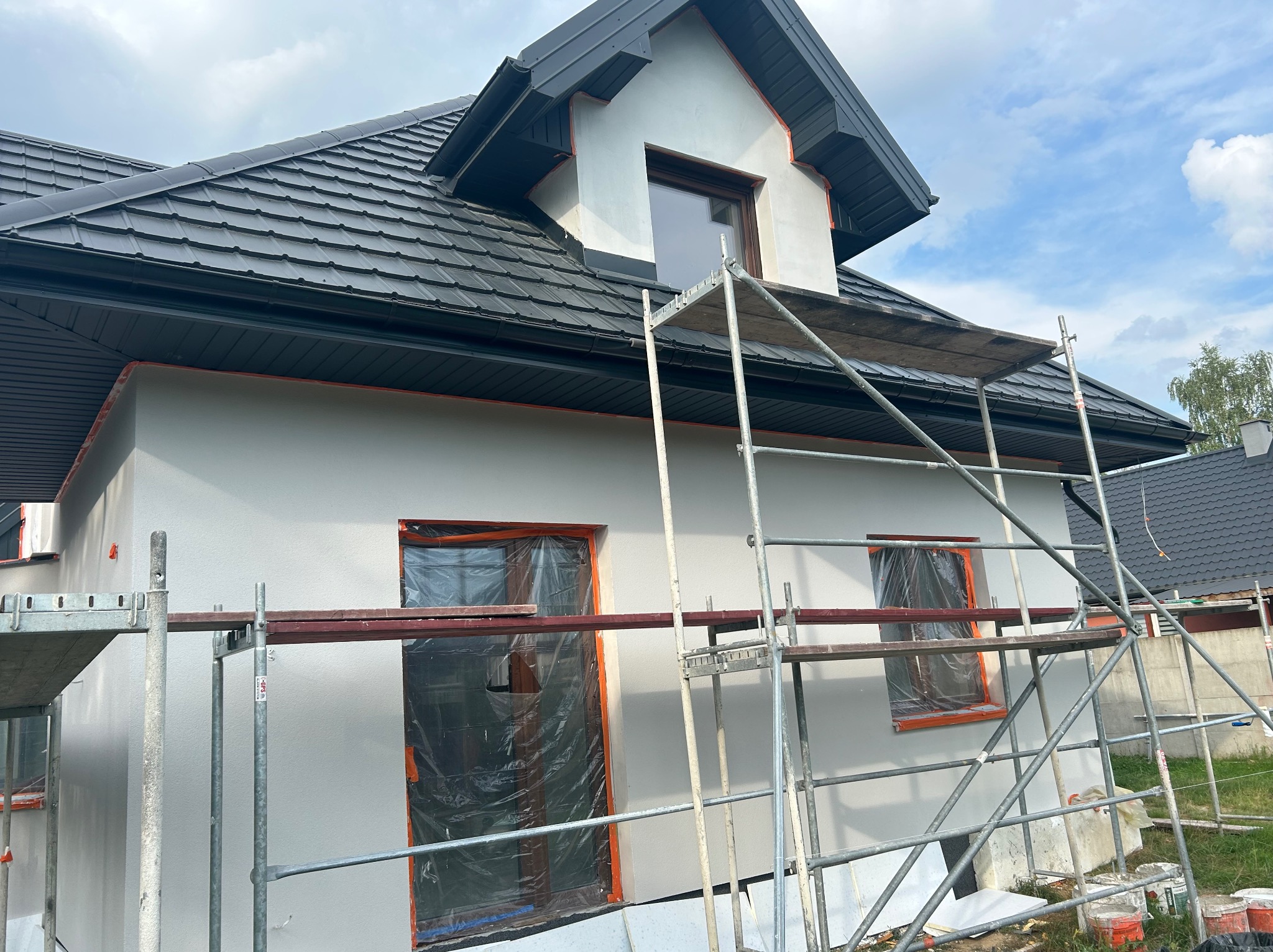 Świeżo otynkowany dom jednorodzinny z czarnym dachem, zabezpieczone okna folią, widoczne rusztowania. Jasne tynki zewnętrzne w trakcie prac wykończeniowych.