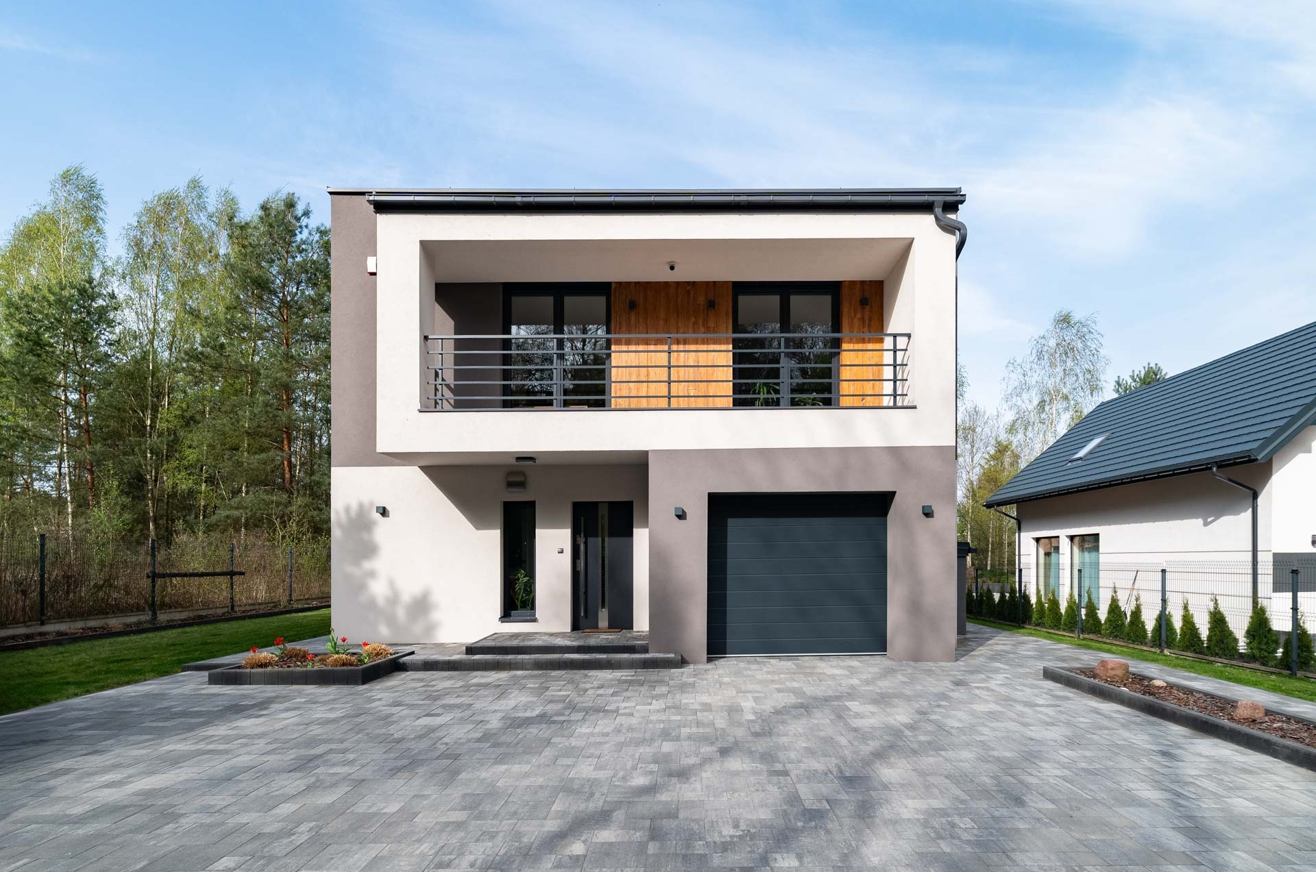 Nowoczesny, dwukondygnacyjny dom z balkonem i garażem, wykończony w bieli i szarości, z brukowanym podjazdem i zadbanym otoczeniem. Minimalistyczny design.