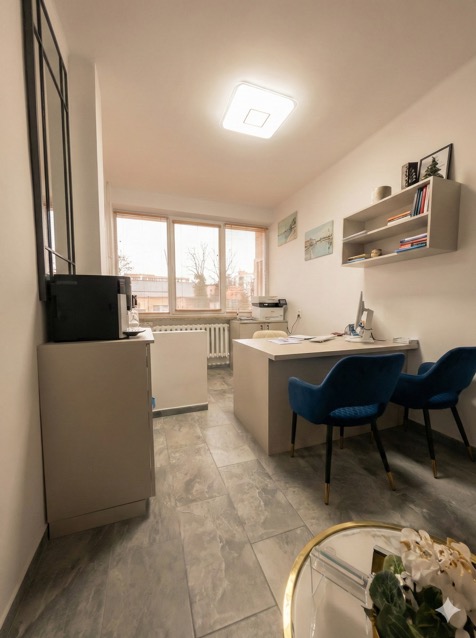 Jasne biuro ubezpieczeń z biurkiem, krzesłami i oknem. Minimalistyczny wystrój z szarymi płytkami podłogowymi i dekoracjami na półce. Widok z perspektywy klienta.