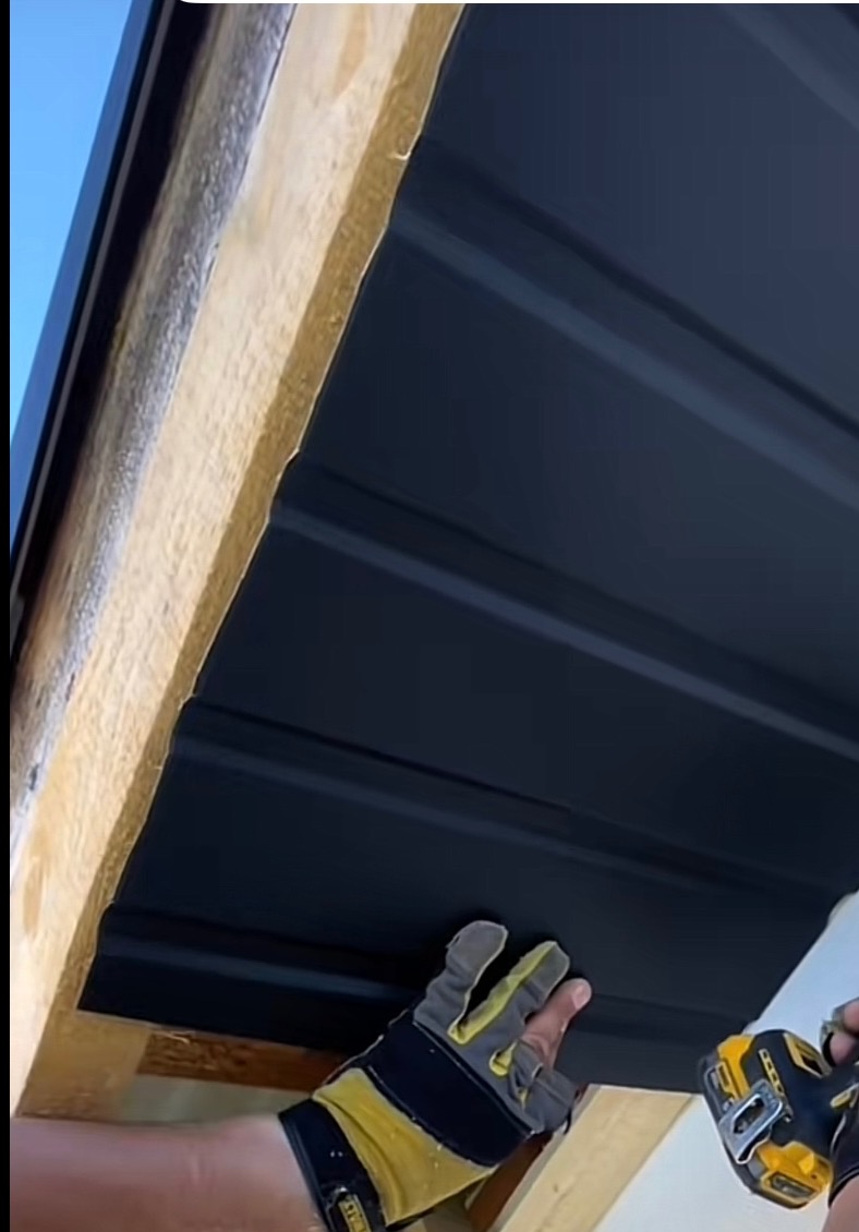 Montaż czarnej blachy trapezowej na drewnianej konstrukcji dachu, widoczne rękawice robocze i wkrętarka DeWalt, ujęcie z dołu.