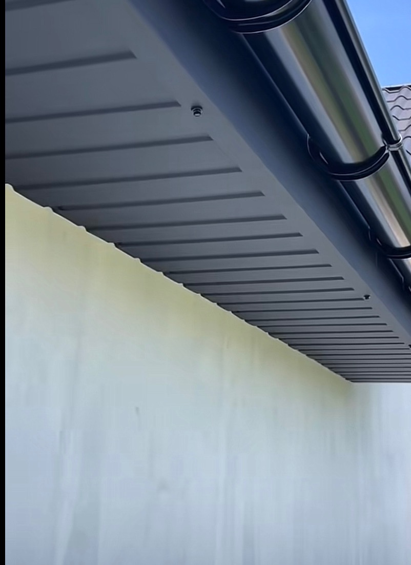 Szary podbitka dachowa z widoczną rynną w kolorze czarnym, zamontowana przy jasnej ścianie budynku. Ujęcie z dołu, podkreślające detale wykończenia.