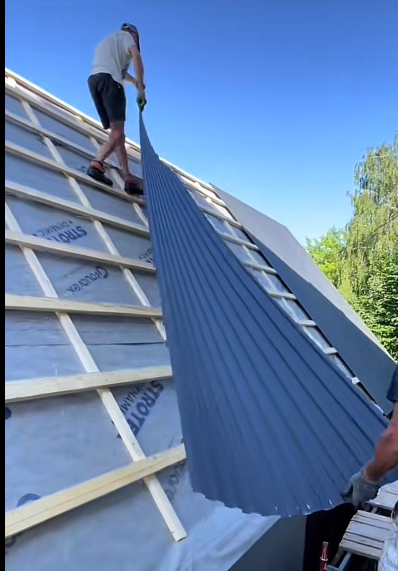 Montaż blachy na dachu z widoczną membraną i łatami. Dekarz w trakcie pracy, trzyma arkusz blachy. Widok z dołu na fragment dachu i niebo.