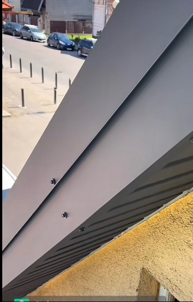 Szary dach z blachy trapezowej, widok z dołu pod kątem, z widocznymi śrubami mocującymi. W tle ulica z zaparkowanymi autami i budynkami.