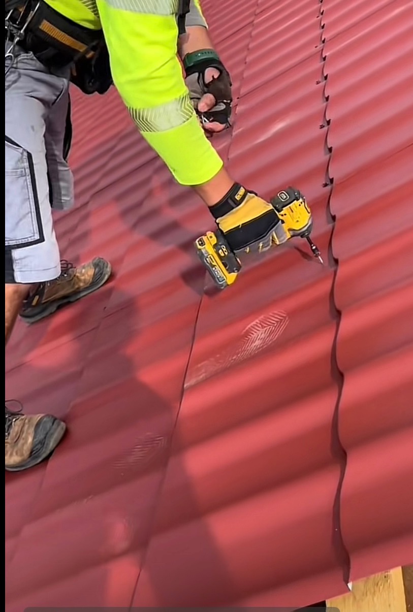 Pracownik w odzieży roboczej mocuje czerwoną blachodachówkę wkrętarką. Widoczne ślady butów na powierzchni dachu. Ujęcie z góry, skupione na narzędziu i dłoniach.