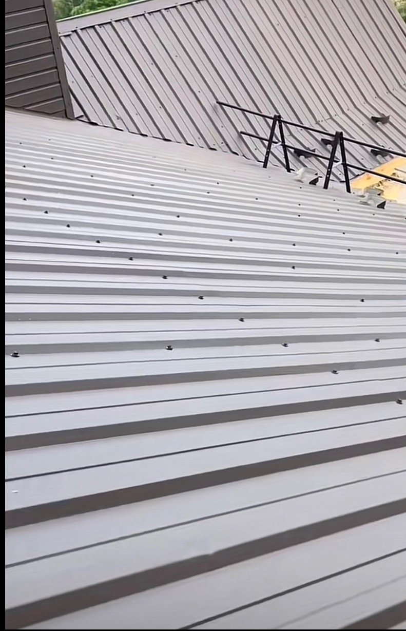 Szary dach z blachy trapezowej, widoczne mocowania i fragment konstrukcji zabezpieczającej na skośnej powierzchni. Ujęcie z dołu, podkreślające regularność wzoru.