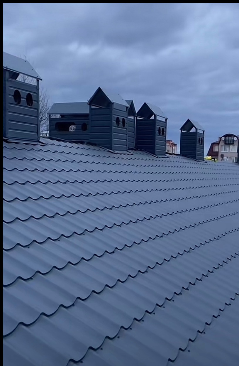 Szary dach z blachy trapezowej z czterema kominami wentylacyjnymi w kształcie domków, widok z dołu na tle pochmurnego nieba.