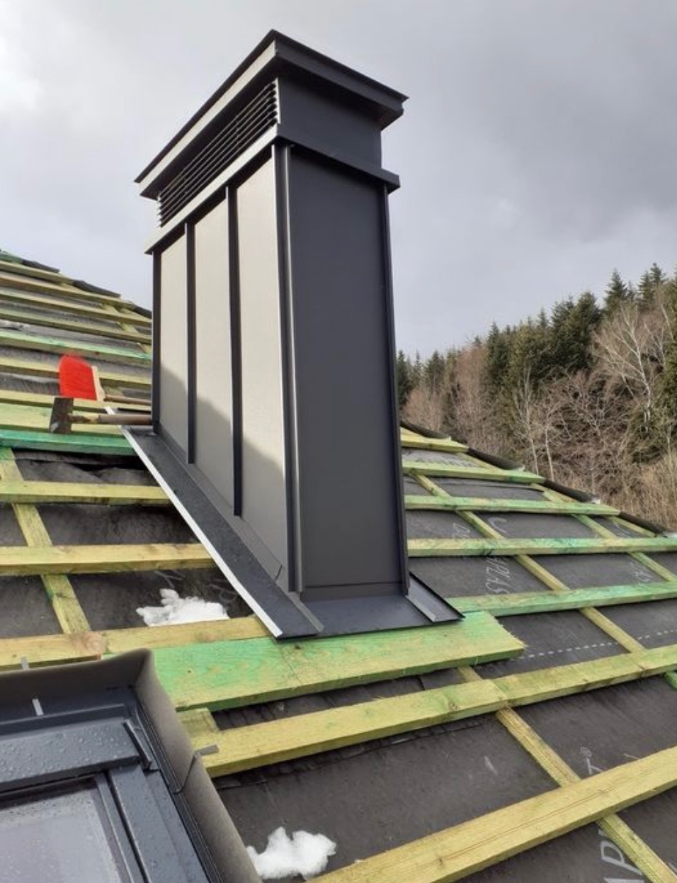 Czarny komin wentylacyjny na dachu w trakcie budowy, widoczne łaty, membrana dachowa i fragment okna dachowego. W tle las i pochmurne niebo.