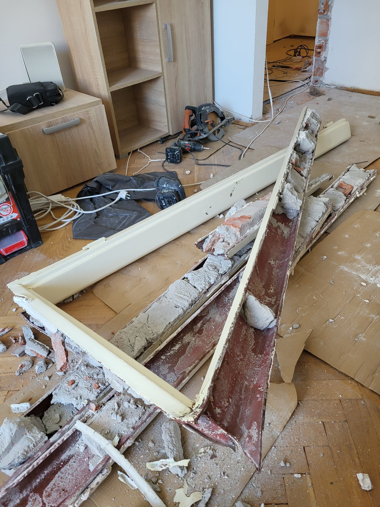 Demontaż ościeżnicy z ceglanego muru. Fragmenty gruzu i narzędzia na podłodze z parkietu. Widoczne ślady po usuniętej futrynie. W tle meble i otwór drzwiowy.