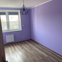 Bud_Job - Pomalowane na fioletowo puste wnętrze pokoju z drewnianą podłogą, białym grzejnikiem i geometryczną lampą wiszącą. Widok zza uchylonych drzwi.