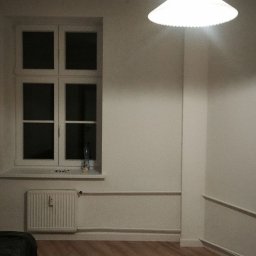 Bud_Job - Pomalowane na biało wnętrze pokoju z drewnianą podłogą, oświetlone lampą sufitową. Widoczne okno z grzejnikiem i dekoracyjnymi listwami przypodłogowymi.