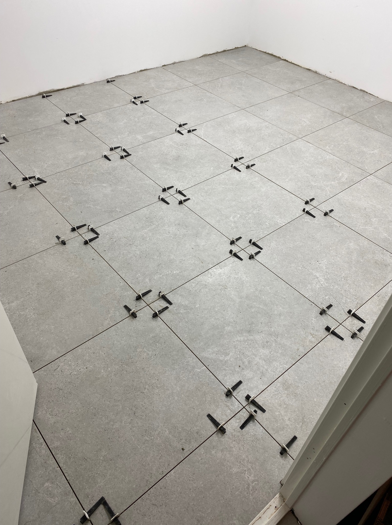 Szare płytki podłogowe w trakcie układania, z czarnymi klipsami poziomującymi w spoinach. Widok z góry, fragment ściany i futryny drzwi.