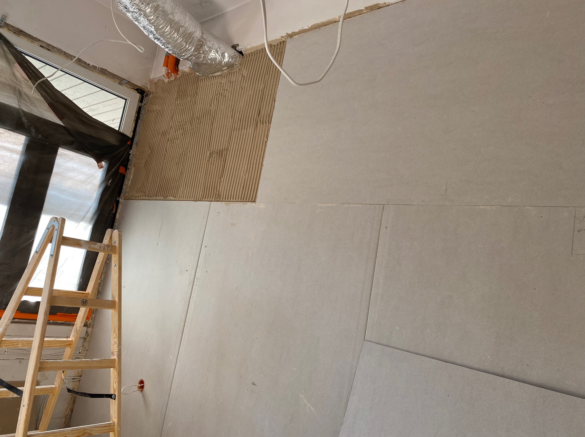 Montaż płyt karton-gips na ścianie z widoczną warstwą kleju, drabina oparta o ścianę, zabezpieczone okno folią. Prace wykończeniowe w trakcie.