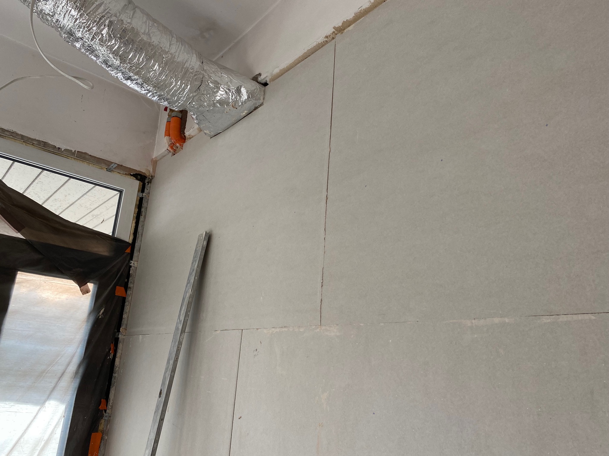 Ściana z płyt karton-gips, widoczne łączenia, częściowo zakryte okno folią ochronną, aluminiowy przewód wentylacyjny pod sufitem. W tle widoczna listwa przypodłogowa.